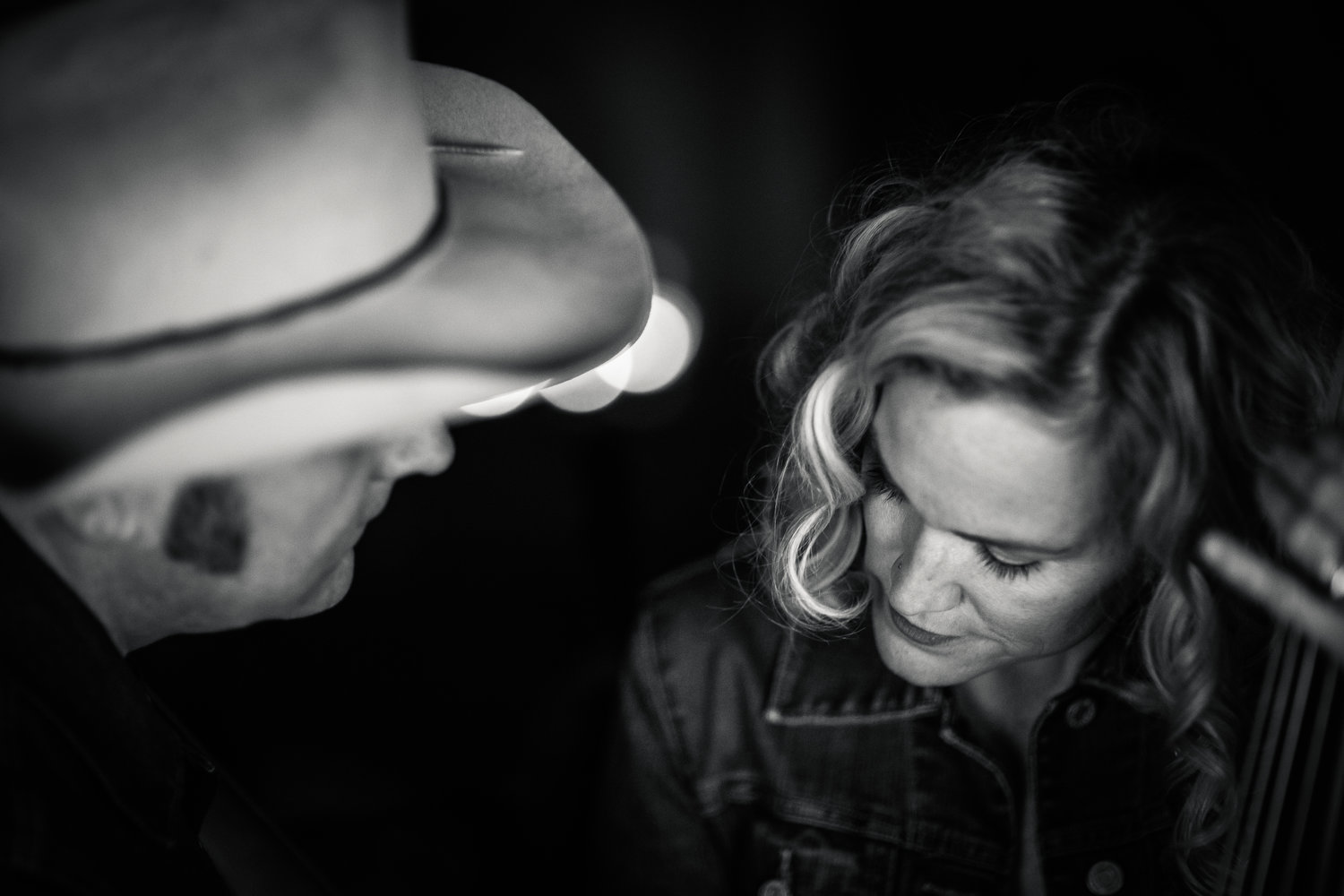 Black and white image of profile of a person wearing a cowboy hat on the left, looking at a woman on the right with shoulder length blonde hair. She is looking down