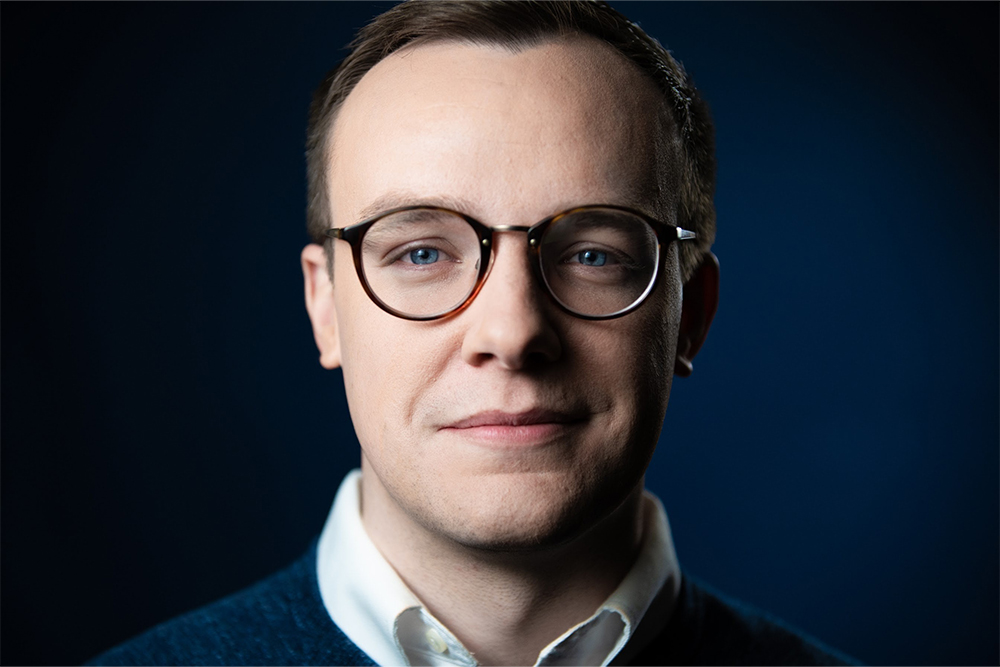 Image of a person with glasses wearing a collared shirt in front of a dark blue background.