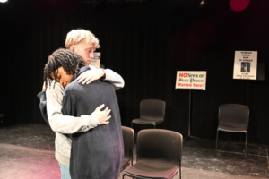 A blond haired young white man comforts a dark-haired young black woman with a hug on a stage with black chairs in and a union protest sign in the background