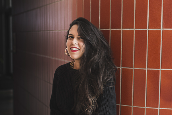 Zaina Alsous smiling at the camera. Her hair is down and she is wearing a black shirt. Alsous is posing in front of a red brick wall.