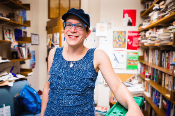 Stephanie Burt standing in her office, smiling at the camera. Burt is wearing a blue tank-top and a black hat.