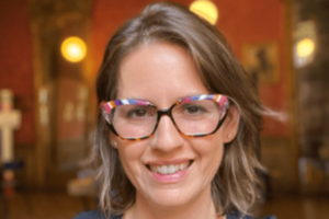 Juliet Guzzetta smiling at the camera. She is wearing wide-rimmed glasses and has shoulder-length brown hair.