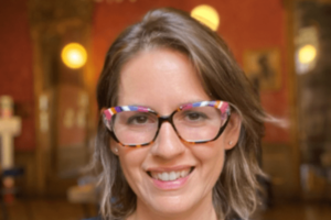 Juliet Guzzetta smiles at the camera. She is wearing wide-rimmed glasses and has shoulder-length brown hair.