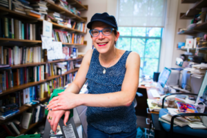 Stephanie Burt standing in their office (a backdrop of bookshelves and books laying around a desk). Stephanie is wearing a blue tank-top and a black hat.