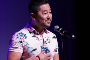 Masaki Takahashi speaking into a microphone on stage. Takahashi has black cropped hair and is wearing a white shirt with a pink and blue floral pattern.