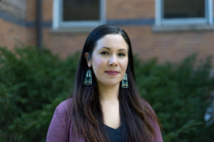 A person with long black hair stands in front of Linton. They are wearing a long-sleeved purple shirt.