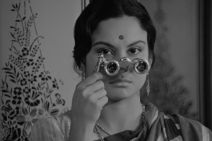 A person with their hair pulled back looks through a small pair of binoculars.