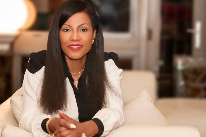 An individual sitting on a white couch. They are wearing a white suite jacket and a black blouse underneath.