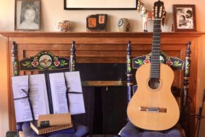 A guitar sitting on a chair next to another chair with music sheets on the seat. Both chairs are situated in front of a fireplace.