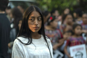 A young person with long, dark hair wears a white shirt and stands in front of a protest.