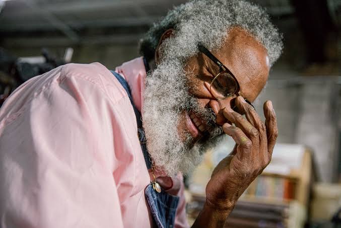 A person wearing a pink shirt, a denim apron, and glasses. They have gray hair and a gray beard. The appear to be laughing in a studio pace.