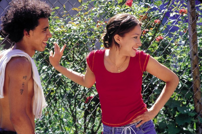 Two individuals standing in a garden. The individual on the right is wearing a red, red short-sleeved shirt. The individual on the left has a white towel draped over their right shoulder.