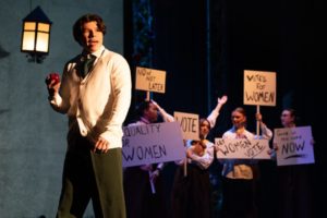 A performer stands beneath a stage lantern holding a red apple, while a group of student performers behind hold protest signs reading “Votes for Women,” “Equality for Women,” “Let Women Vote,” “Now Not Later,” and “Give Us the Vote Now.”