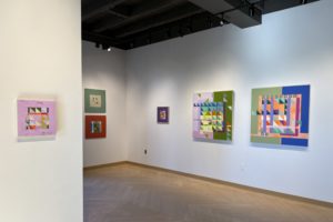 Gallery interior featuring several abstract geometric paintings in pastel tones of pink, green, blue, and purple. The artworks hang on white walls under ceiling track lighting, with wood-patterned flooring below.