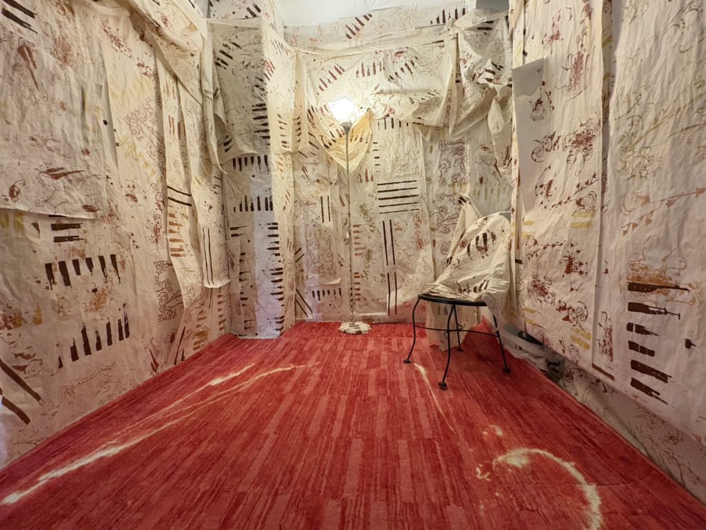 Installation view of a small room with walls and ceiling draped in cream-colored fabric marked with brown and red abstract prints and linear patterns. The floor is covered in red textured material, and a single chair sits near the back wall under a hanging light.