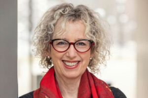 A professional headshot of a woman with short, curly blonde hair and glasses with red frames. She is smiling and wearing a red patterned scarf over a dark top, positioned against a brightly lit, blurred indoor background.
