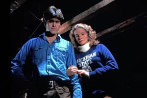 A dramatic, low-angle cinematic still. A man in a blue button-down shirt stands in a dimly lit, rustic interior with heavy wooden ceiling beams. To his right, a woman with blonde hair holds his arm; she is wearing a blue sweatshirt prominently featuring the "Michigan State" name.