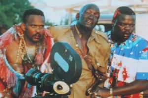 Three men gathered closely behind a professional camera. The individual on the left wears a feathered garment, the man in the center is in a khaki uniform, and the man on the right wears a vibrant, patterned shirt with red stains.