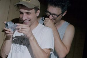 A candid, low-light photograph of two men indoors. The man in the foreground wears a tan baseball cap and a white t-shirt with a line-art graphic; he is smiling broadly while looking down at a small, handheld silver digital camcorder in his hands. Behind him, a second man with dark curly hair and glasses leans in, laughing.
