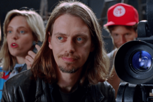 A close-up production still featuring three individuals behind a professional film camera. In the center, a man with long brown hair and a goatee looks off-camera. To his left, a woman with blonde hair with an open-mouthed expression. In the background, a man wears a bright red baseball cap.
