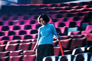 A woman with short dark hair stands in the center of a large, empty theater with rows of unoccupied red velvet seats rising behind her. She wears a light blue turtleneck and a dark patterned skirt, holding a long, thin red pole or staff. The lighting is moody.