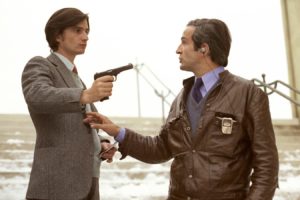 Two men standing on outdoor concrete steps. The man on the left, wearing a grey blazer, holds a small handgun leveled toward the man on the right. The man on the right wears a brown leather jacket and has an earpiece in his ear, looking at the other man with a serious expression. Light snow is visible on the steps in the background.
