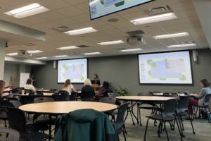 A classroom or presentation hall with round tables and chairs where several attendees watch a speaker presenting slides on two large projection screens.