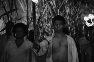 A black and white image showing a group of people standing at night in front of tall, dense vegetation resembling sugarcane. In the center, a Black man with an intense, piercing stare holds a flaming wooden torch aloft in his right hand. He is wearing a light-colored, unbuttoned shirt that exposes his chest. To his left stands a Black woman with dark, textured hair, wearing a light-colored v-neck top. Other figures are partially visible in the shadows behind them, and another flaming torch can be seen held by someone on the far right edge of the frame. The scene is dramatically illuminated by the firelight.