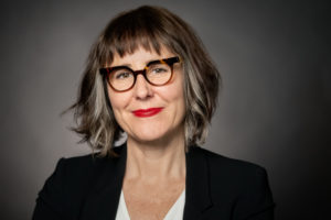 Woman wearing a black blazer, glasses, and red lipstick smiling at the camera