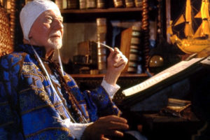 An elderly man with a white goatee, wearing a white cap tied under his chin and an ornate blue and gold patterned robe, sits at a slanted writing desk. He holds a quill pen in his raised hand and is looking thoughtfully upward and away from the document in front of him. The warmly lit background reveals a study filled with bookshelves, a small globe, and a model of a sailing ship