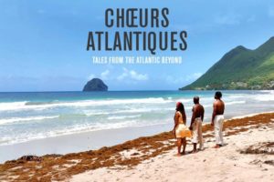 A promotional image featuring the text 'CHŒURS ATLANTIQUES' in large, dark grey letters, with the subtitle 'TALES FROM THE ATLANTIC BEYOND' in smaller white letters below it. The background is a scenic beach where three people—a woman in a yellow dress with a red headwrap and two shirtless men in light-colored pants—stand on the sand looking out at the ocean. A line of brown seaweed stretches along the shoreline. The bright blue water features gentle white waves, and in the distance, there is a large, rounded rock island in the ocean and a steep, lush green mountain rising from the coastline on the right under a partly cloudy sky.
