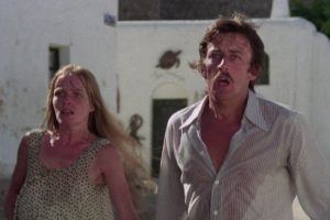 A woman with long blonde hair in a sleeveless patterned top and a man with a mustache in a lightly striped, partially unbuttoned shirt stand outdoors. Both have wide-eyed expressions of shock and fear as they look off-camera to the left. In the background are white stucco buildings with an arched doorway