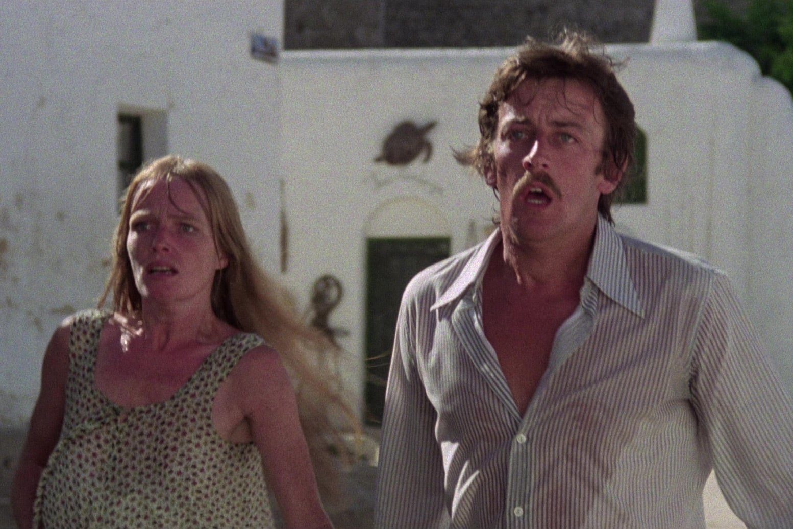 A woman with long blonde hair in a sleeveless patterned top and a man with a mustache in a lightly striped, partially unbuttoned shirt stand outdoors. Both have wide-eyed expressions of shock and fear as they look off-camera to the left. In the background are white stucco buildings with an arched doorway