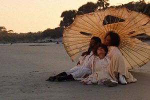 Three people—an older woman and two children—sit close together on a sandy beach, sheltered beneath a large, tattered paper parasol. They are wearing vintage, light-colored, long-sleeved clothing. The group is looking thoughtfully off to the left. The scene is bathed in the warm, golden light of sunset or sunrise, with a dark, tree-lined shoreline visible in the background