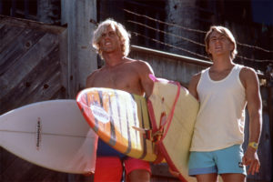 Two young men stand holding surfboards in bright sunlight in front of a wooden structure topped with barbed wire. The man on the left is shirtless with blonde hair, carrying a white surfboard and an orange-and-yellow striped surfboard. The man on the right wears a white tank top and light blue shorts, holding a yellow surfboard. Both men are looking upward and squinting slightly.