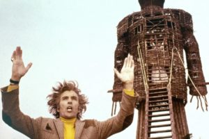 A man with messy brown hair, wearing a yellow turtleneck under a brown patterned jacket, stands in the foreground with his arms raised high and his mouth open as if shouting or chanting. Behind him looms a massive, towering human-shaped structure made of woven wicker. Inside a caged section within the chest cavity of the wicker giant, a figure is visible. The scene is set against a pale, overcast sky.